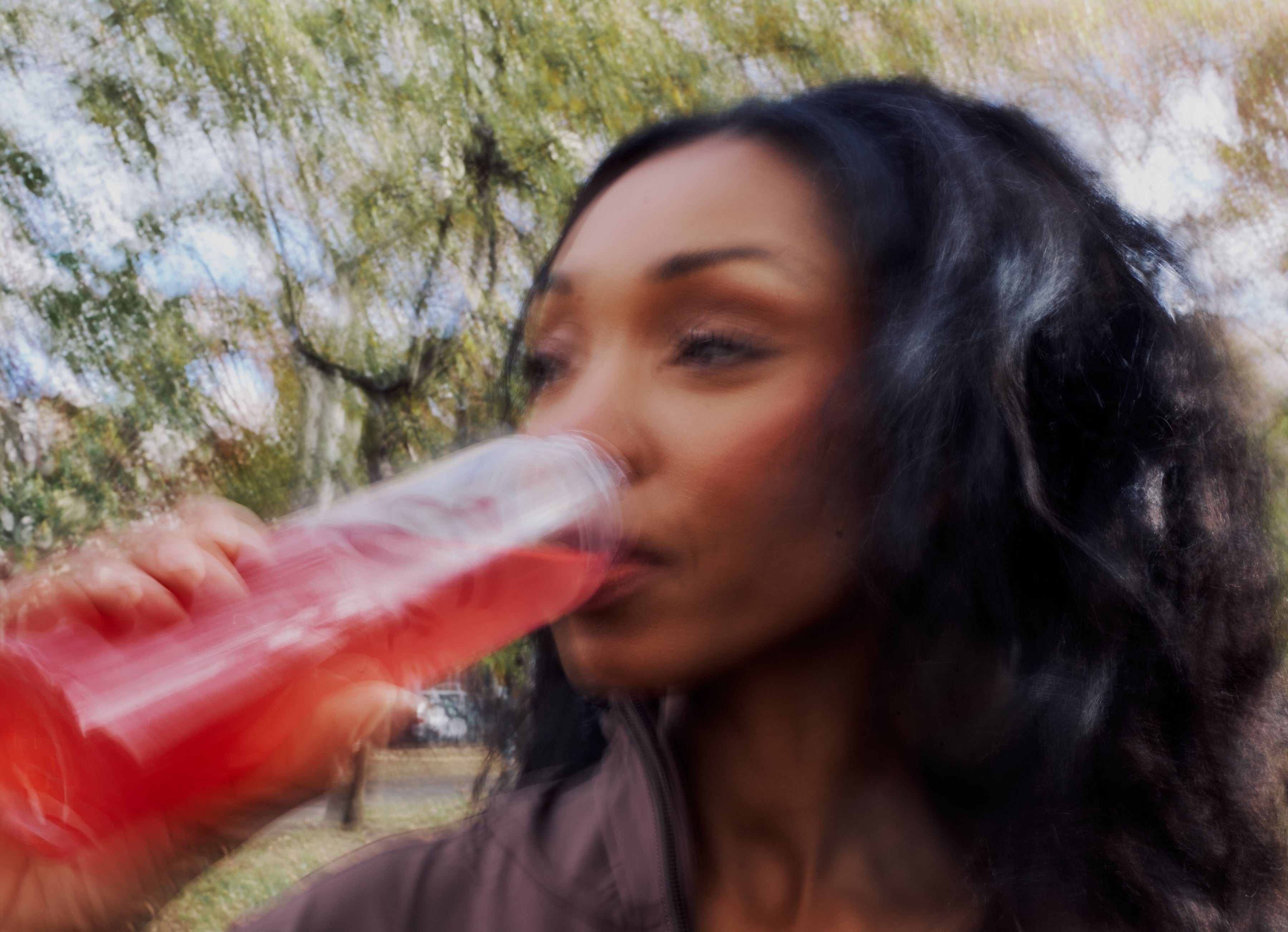 WClose-up of a woman drinking a red ByEla supplement outdoors, capturing movement and everyday vitality.